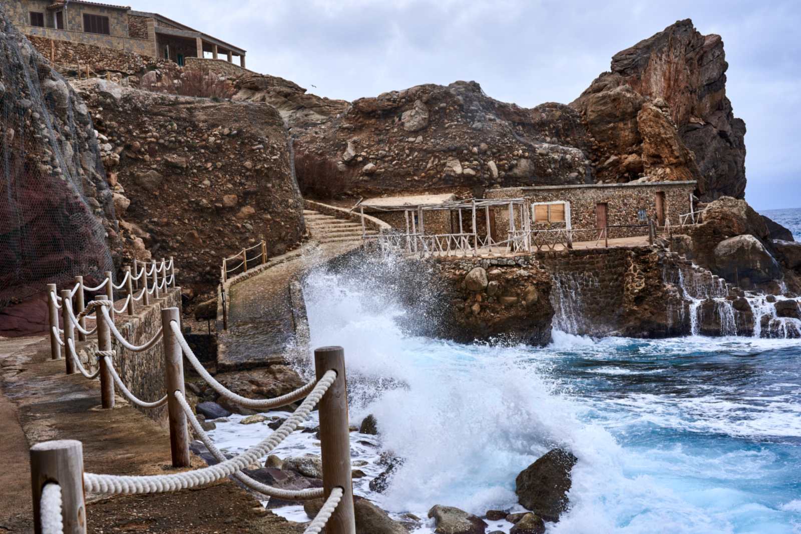 Waves crashing against a rocky Mediterranean shoreline below stone buildings at Cala d'Estellencs, Mallorca