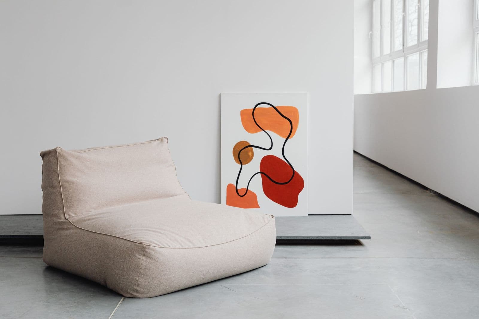 A bold orange and red abstract canvas leaning against a white wall beside a beige lounge chair in a bright, gallery-like room