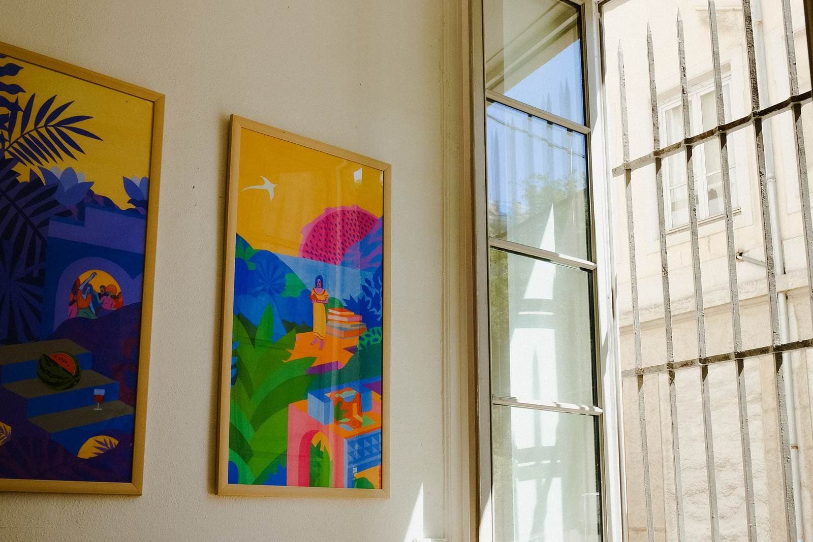Two colorful artworks hanging on a wall beside a tall window in direct sunlight.
