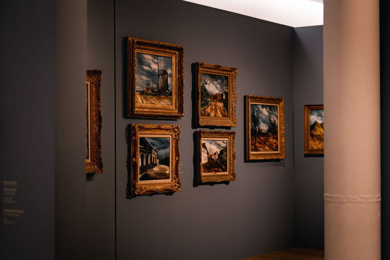 A dim art gallery wall hung with landscape paintings in ornate gilt frames