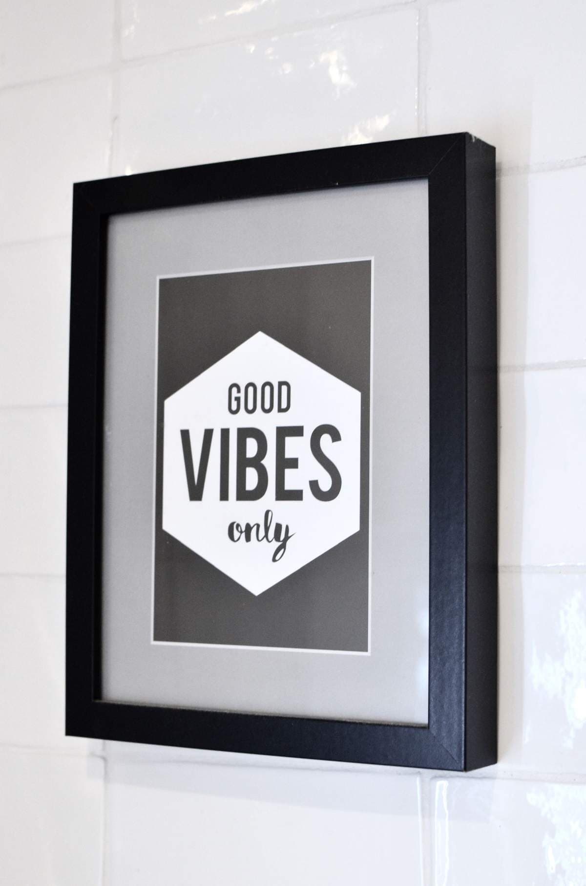 Framed black-and-white typographic print reading Good Vibes Only hanging on a white tiled wall
