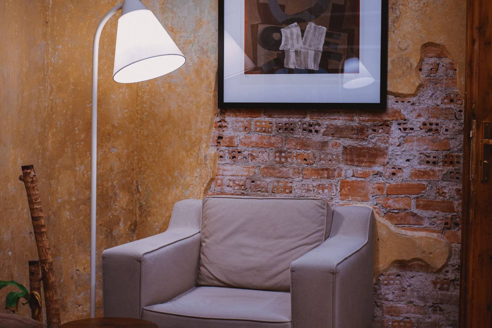 A framed abstract print hanging on an exposed brick and plaster wall above a beige armchair, beside a white floor lamp.