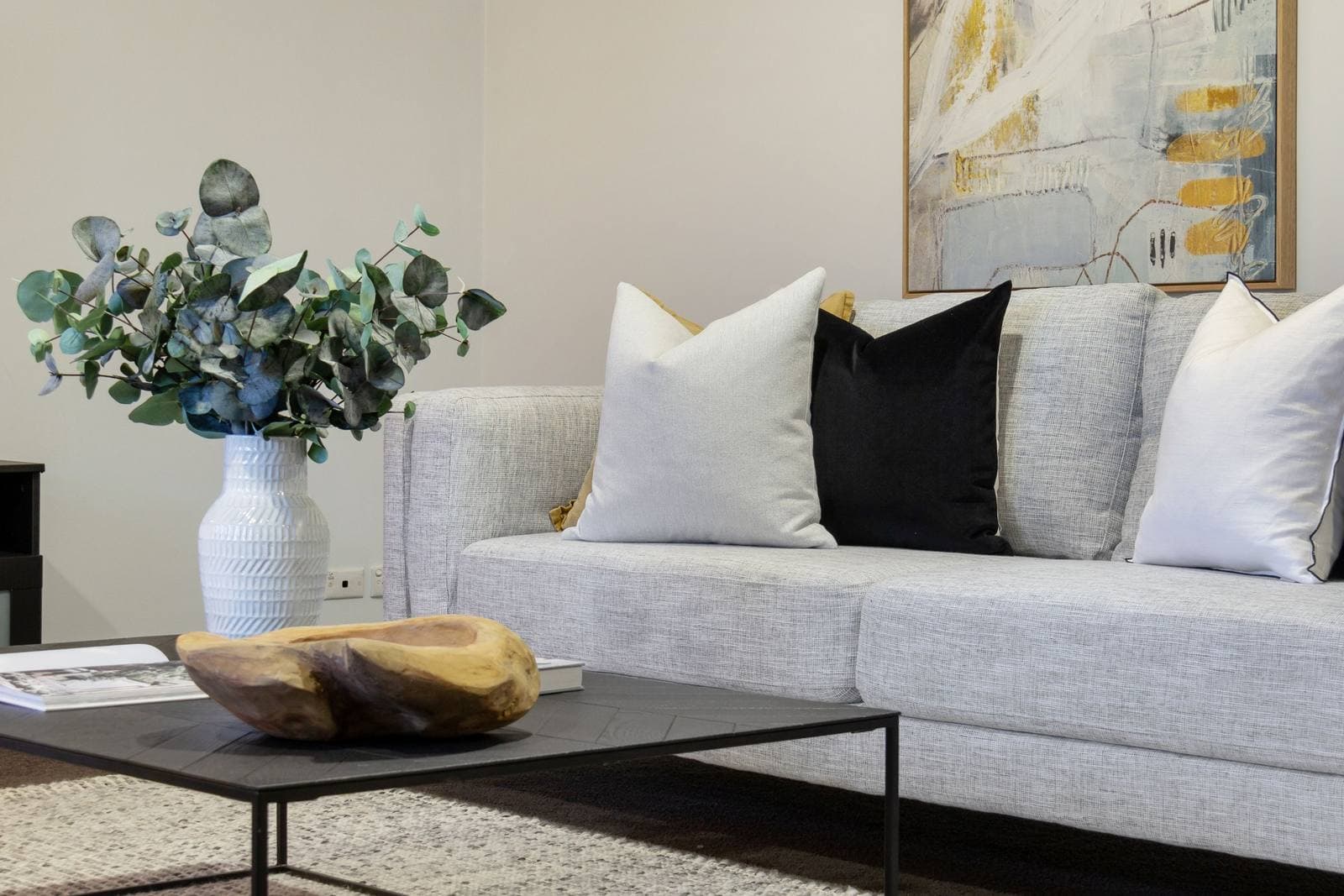 A staged living room with a grey sofa, styled cushions and a large neutral abstract painting above it.