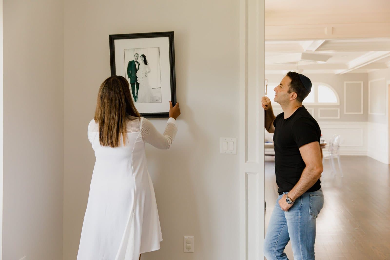 A woman holding a framed wedding photograph against a wall at eye level while a man steps back to check its height and position.
