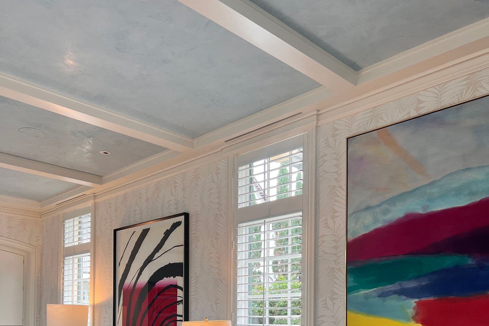 A bright dining room where two colorful abstract paintings — one in black, red and white, the other in sweeping bands of teal, red and gold — bring strong color to pale walls.