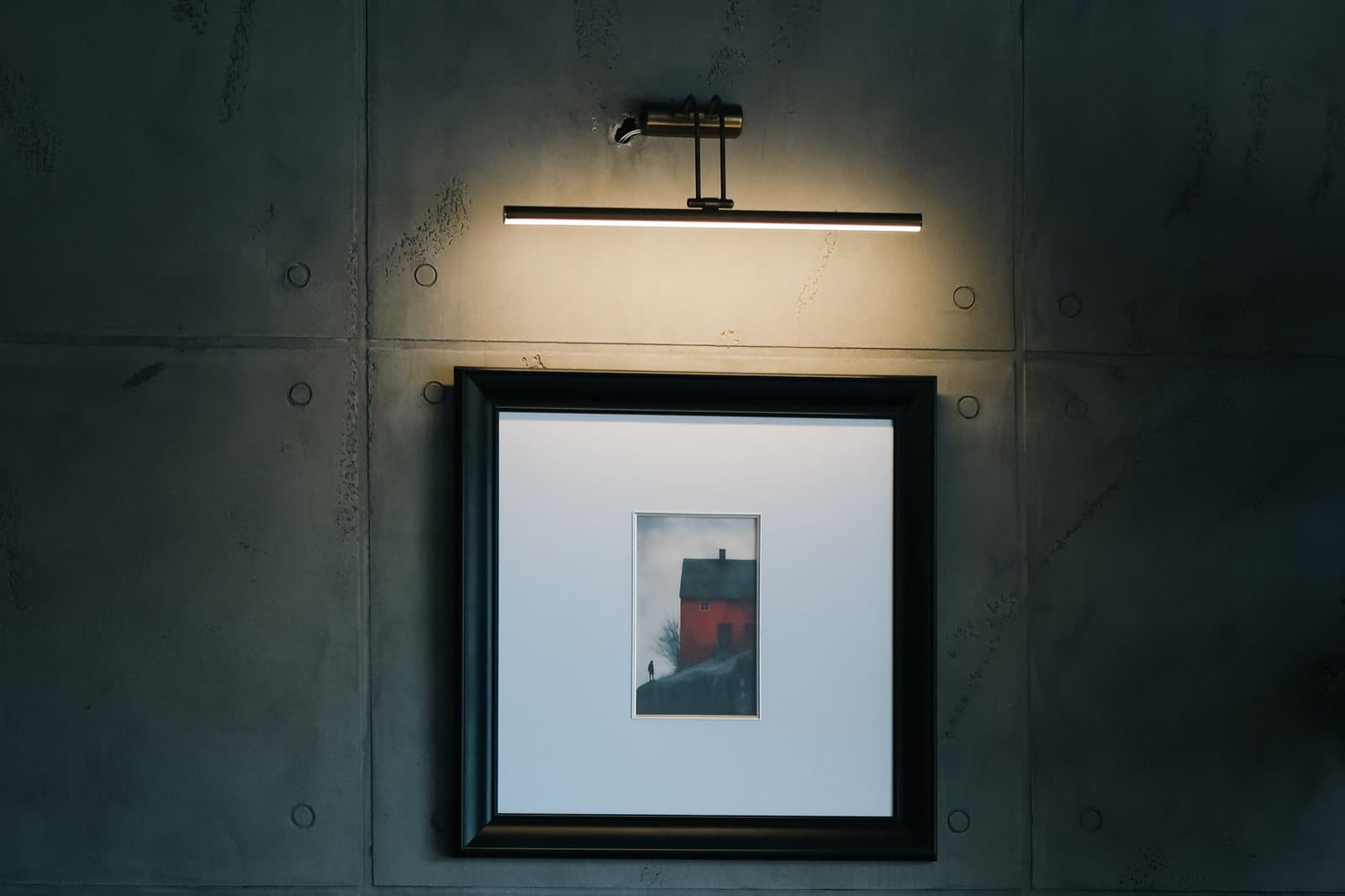 A slim brass picture light mounted above a framed photograph, washing the artwork in warm light on a dark concrete panel wall.