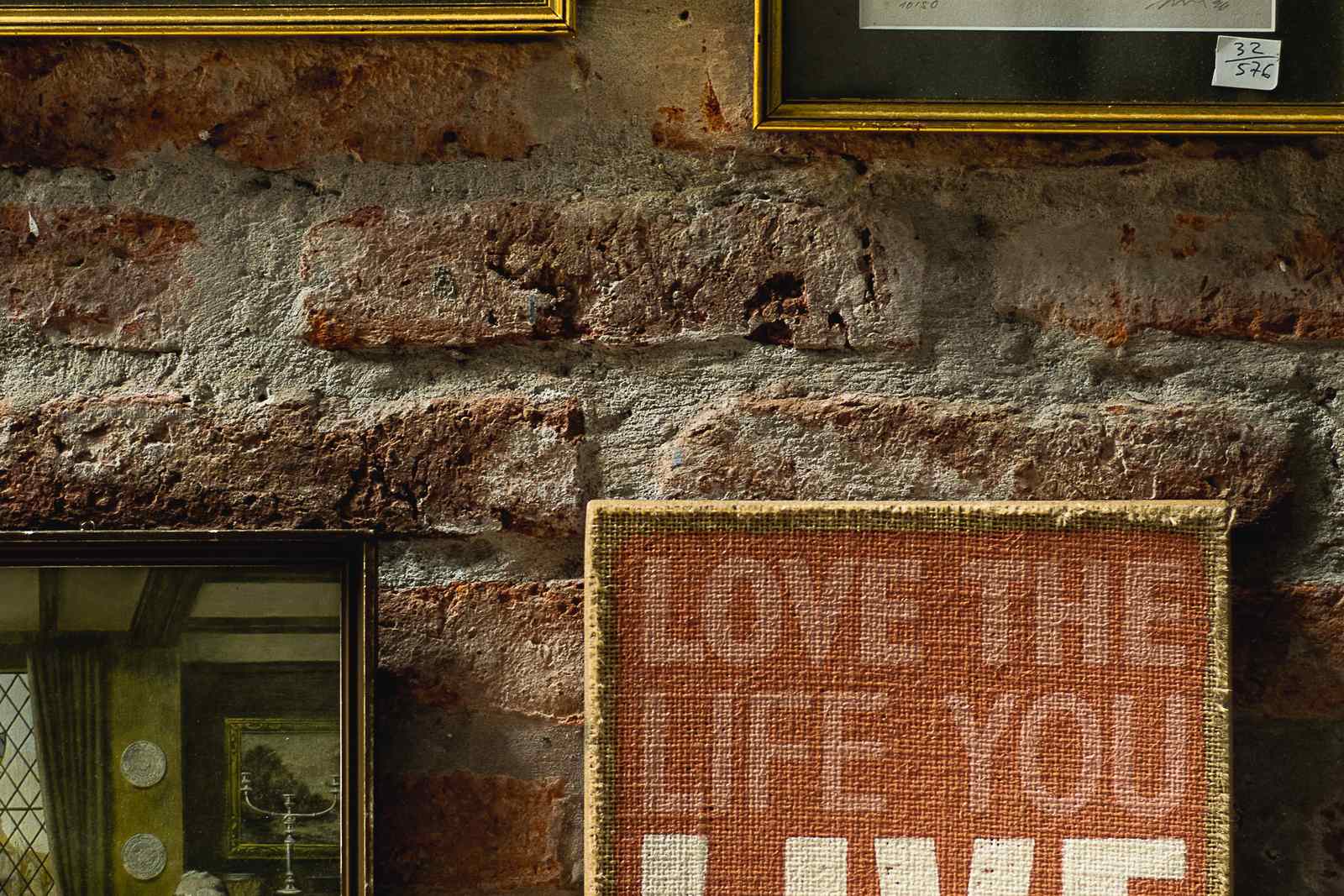 An exposed brick wall mixing gold-framed city prints with a burlap canvas sign reading 'LOVE THE LIFE YOU LIVE' in an eclectic arrangement.