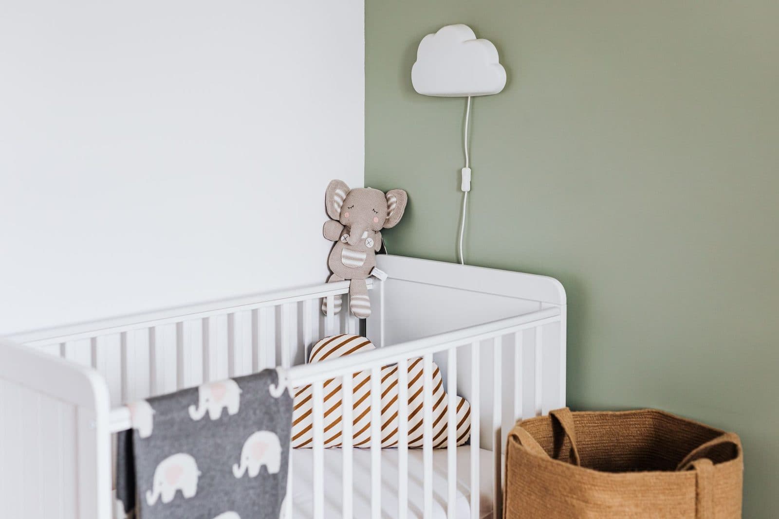Nursery with a white crib against a sage-green wall, a cloud-shaped wall light and a plush elephant on the rail.