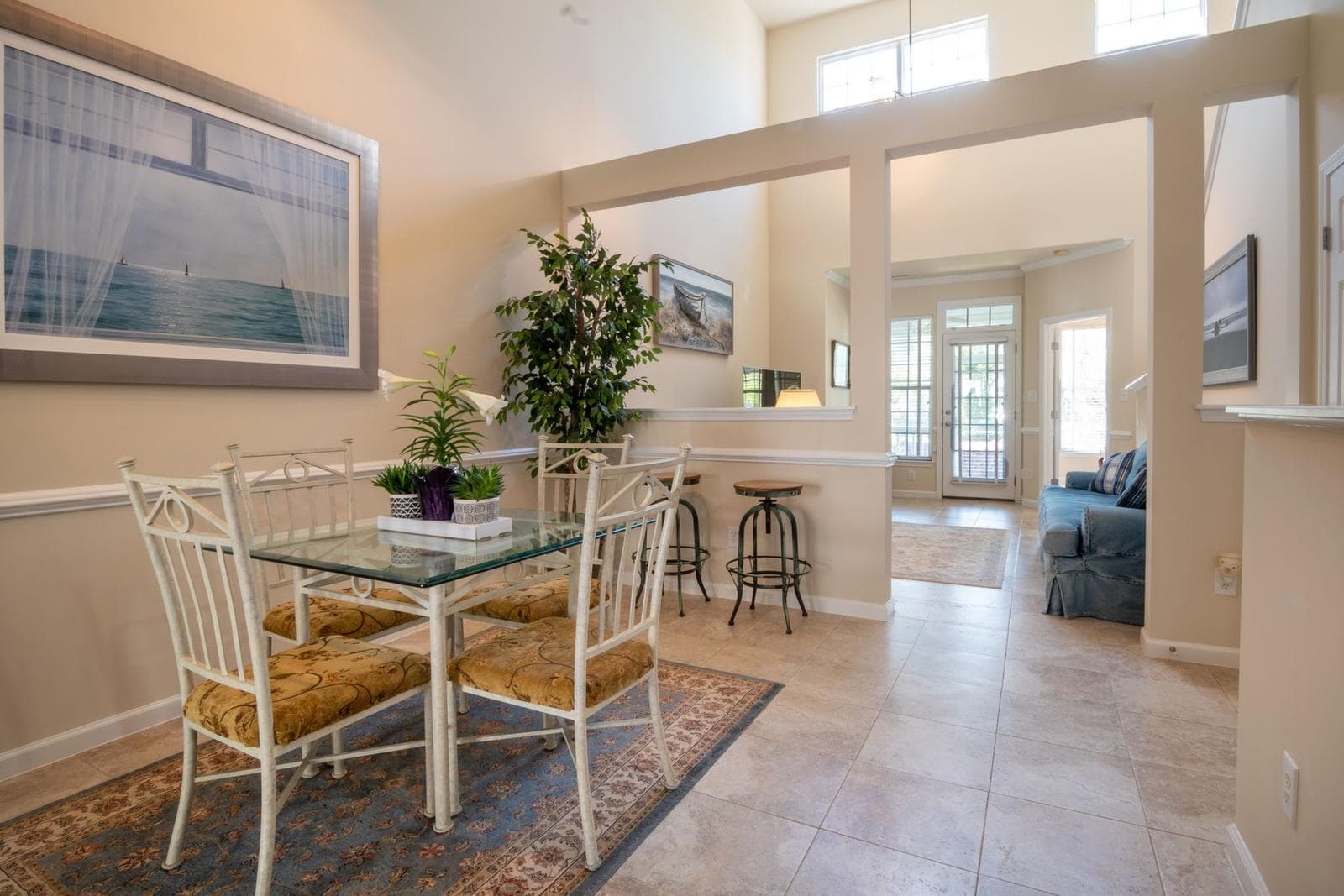 Open-plan dining area with a glass-top table beneath a large framed landscape, with the living space visible through a wide opening beyond.