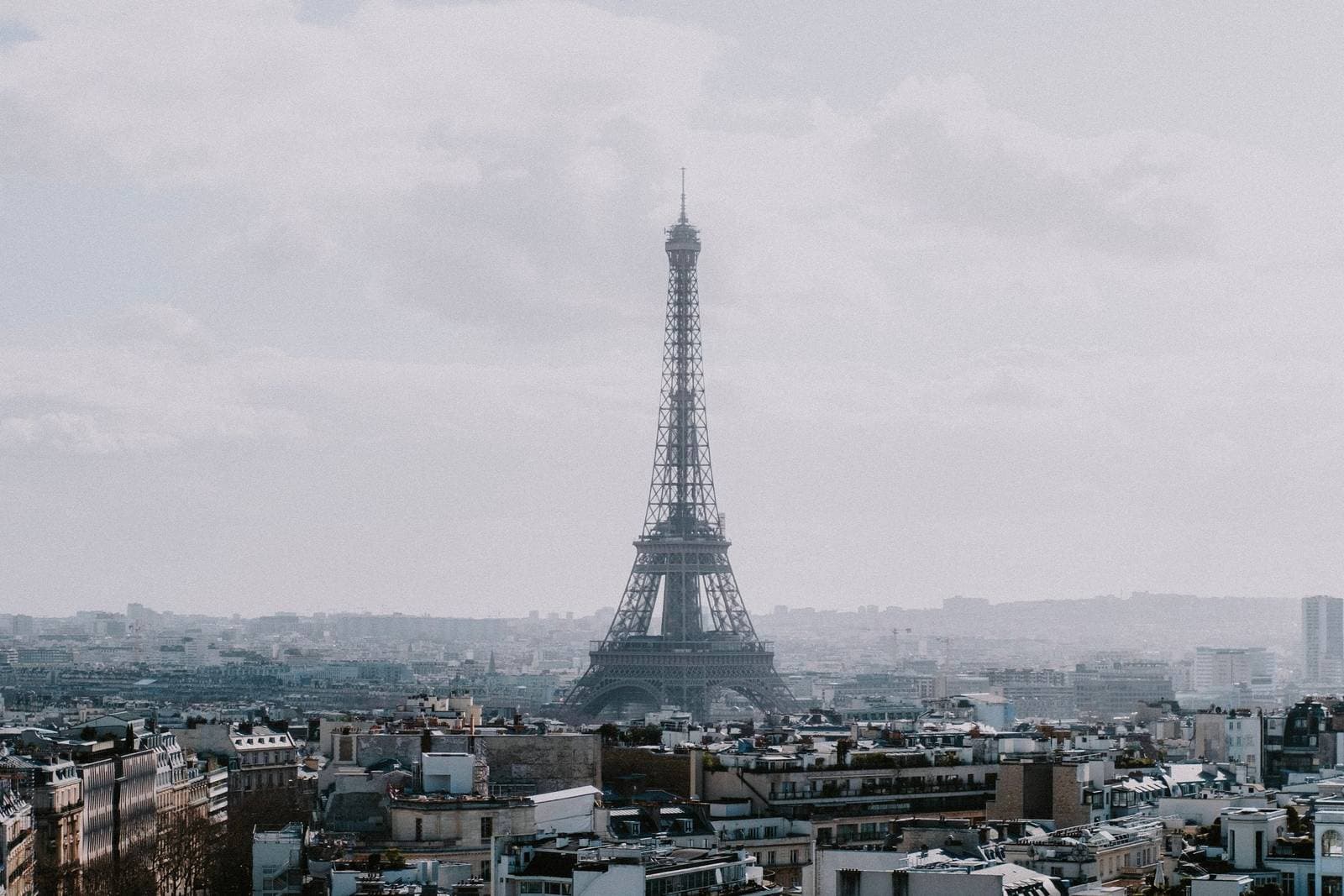 The Eiffel Tower rising above Paris rooftops under a pale, cloudy sky.