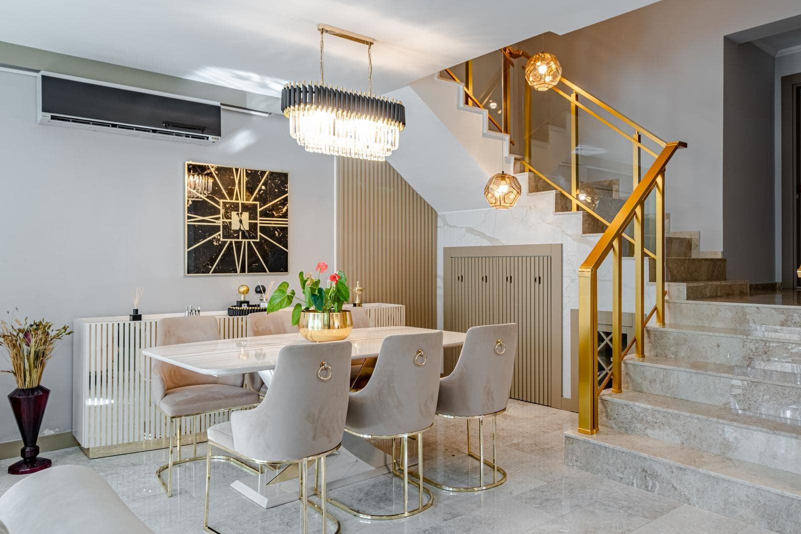Marble staircase with a brass railing rising beside a dining table, with a gold sunburst artwork on the wall opposite.