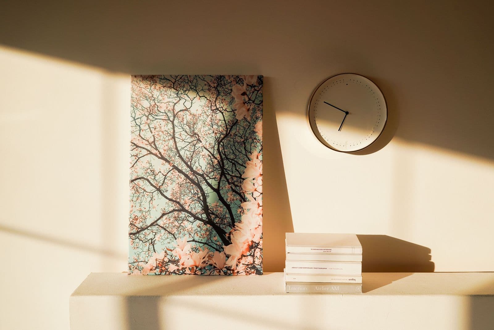 A floral canvas print in soft beige and blush tones leaning on a shelf beside a minimalist wall clock, lit by warm sunlight against a neutral wall.