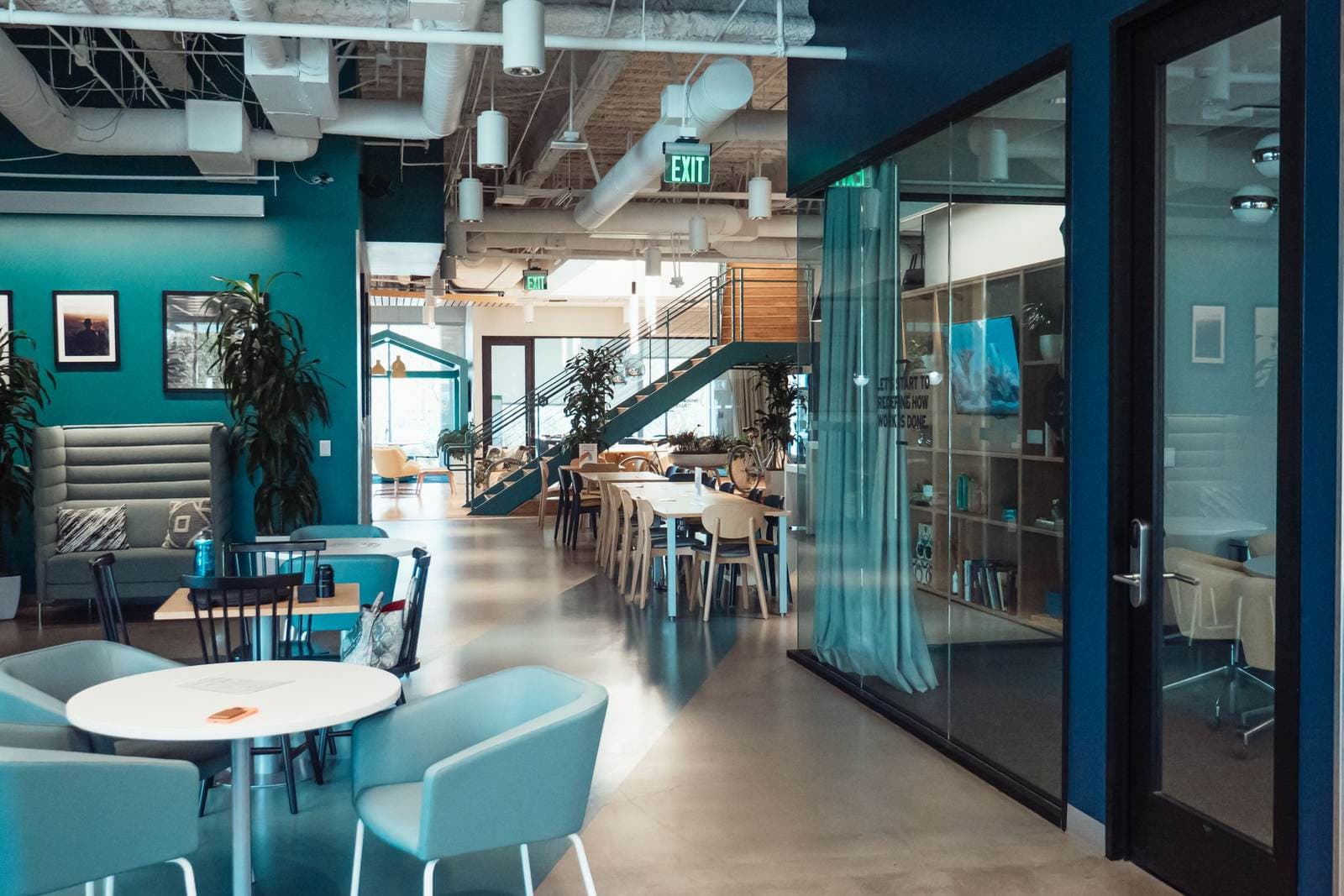 A coworking office lounge with teal walls, cafe tables and framed pictures on the far wall.