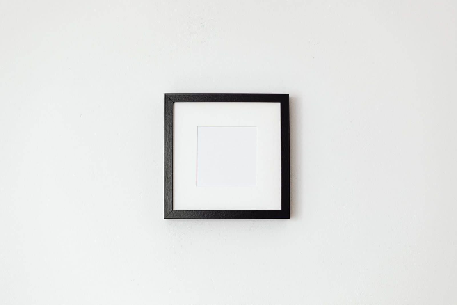 A single small empty black frame hanging alone in the middle of a large blank white wall, far too small for the space around it.