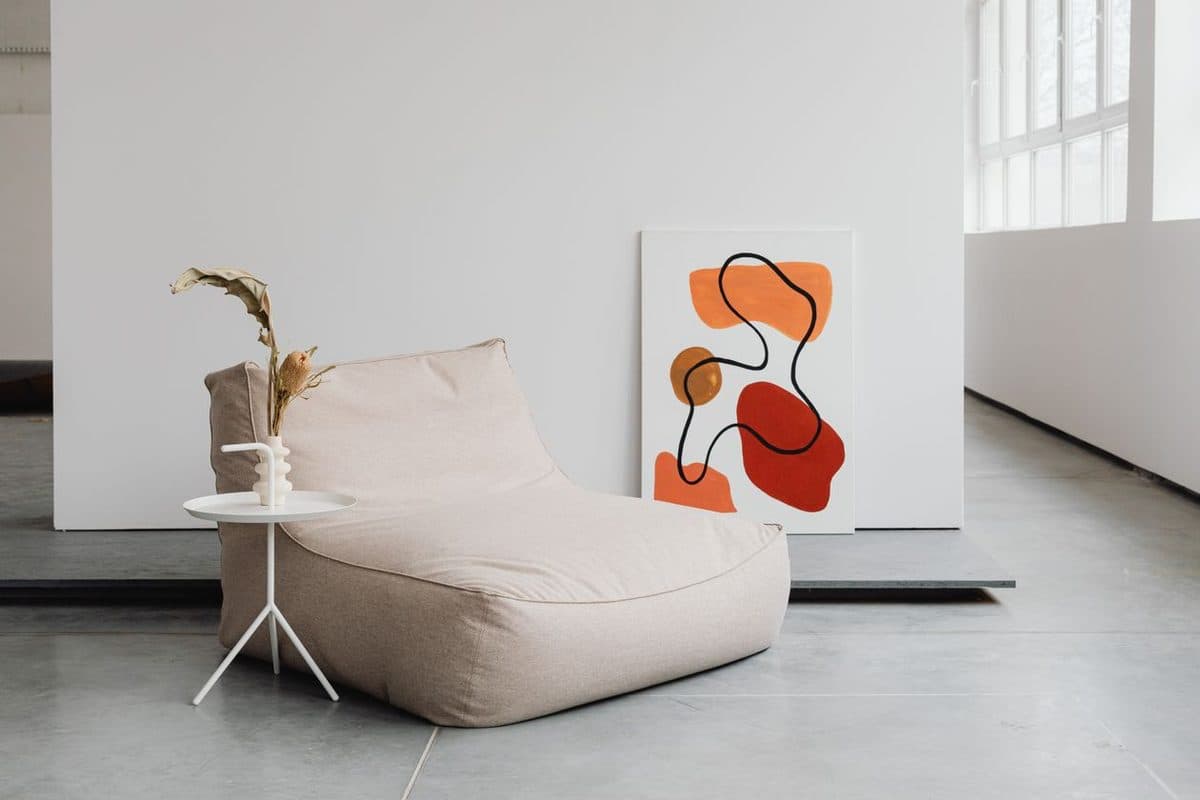 A bold orange and red abstract canvas leaning against a white wall in a neutral minimalist room beside a beige lounge chair