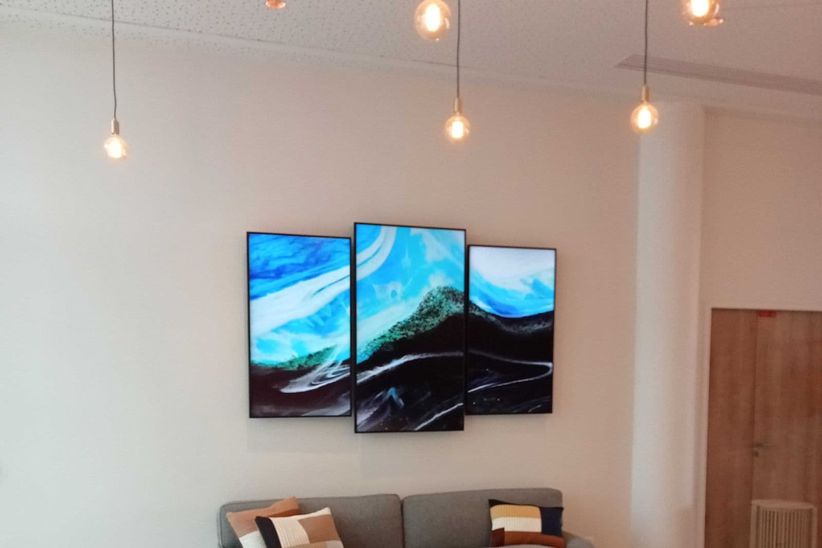Three glossy frameless panels printed with a blue wave design mounted as a triptych on a white wall above a grey sofa in a bright room with pendant lights.