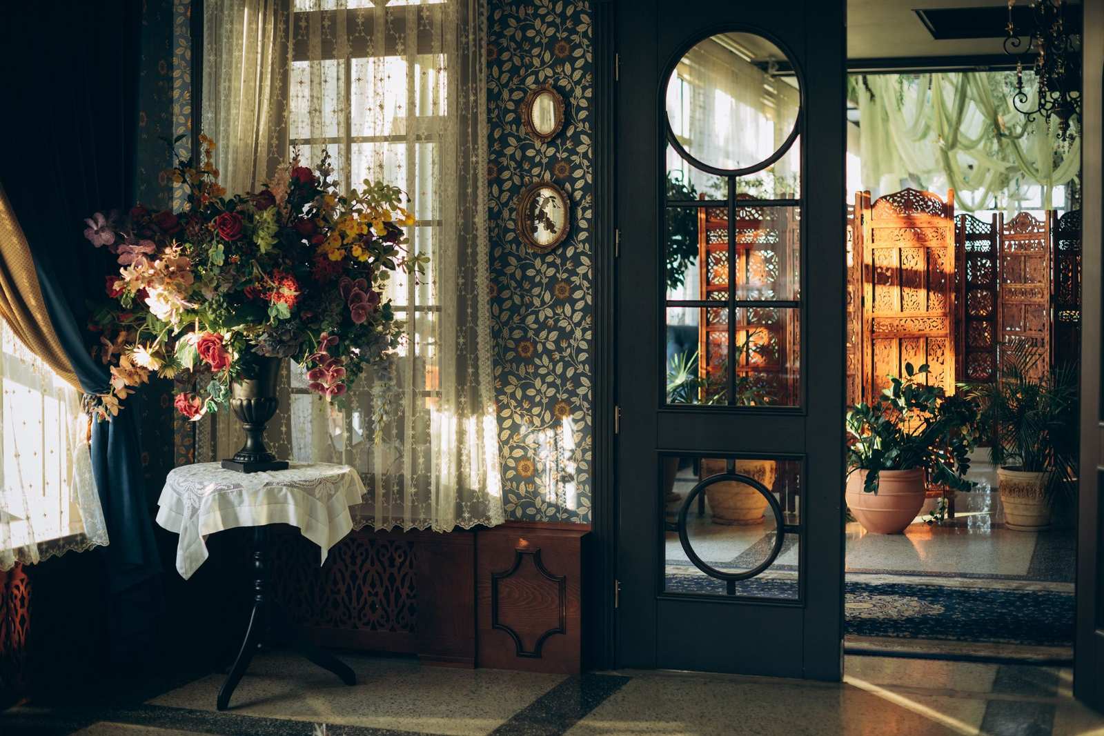 A cottage corner with floral wallpaper, lace curtains, two small oval framed pictures and a large bouquet of garden flowers on a lace-covered table