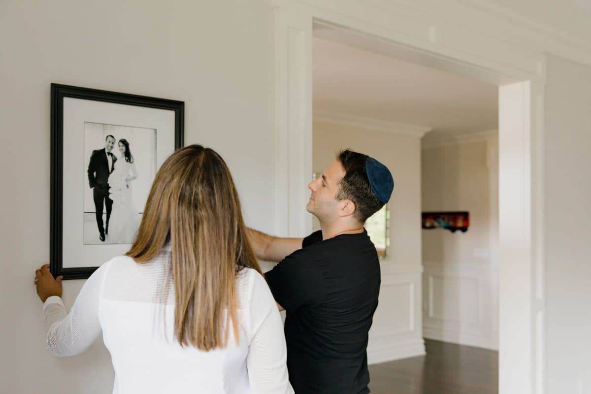 A man and a woman together holding a large framed black-and-white wedding photograph against a white wall