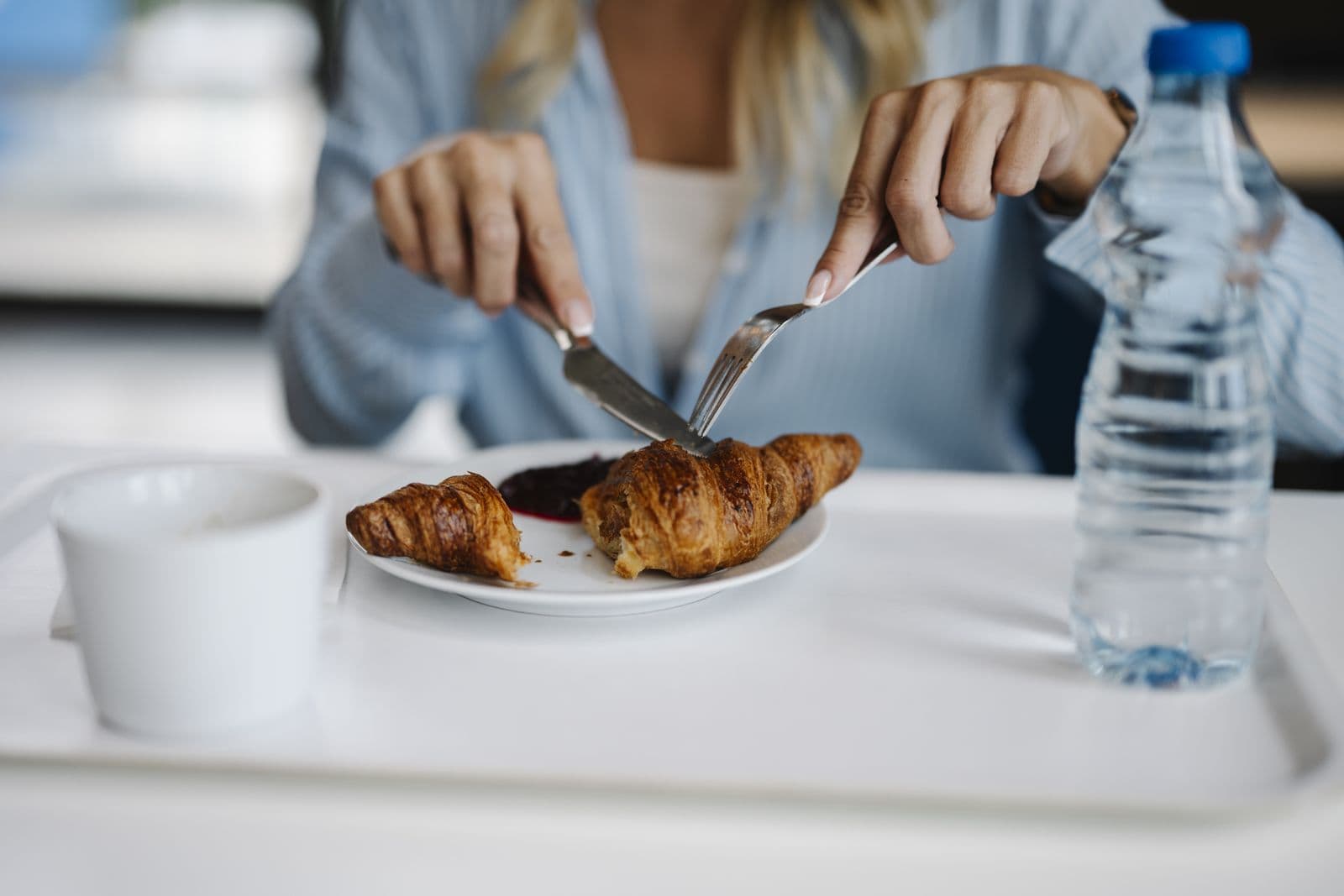 Close-up of a croissant being cut with a knife and fork on a café tray, beside a cup of coffee