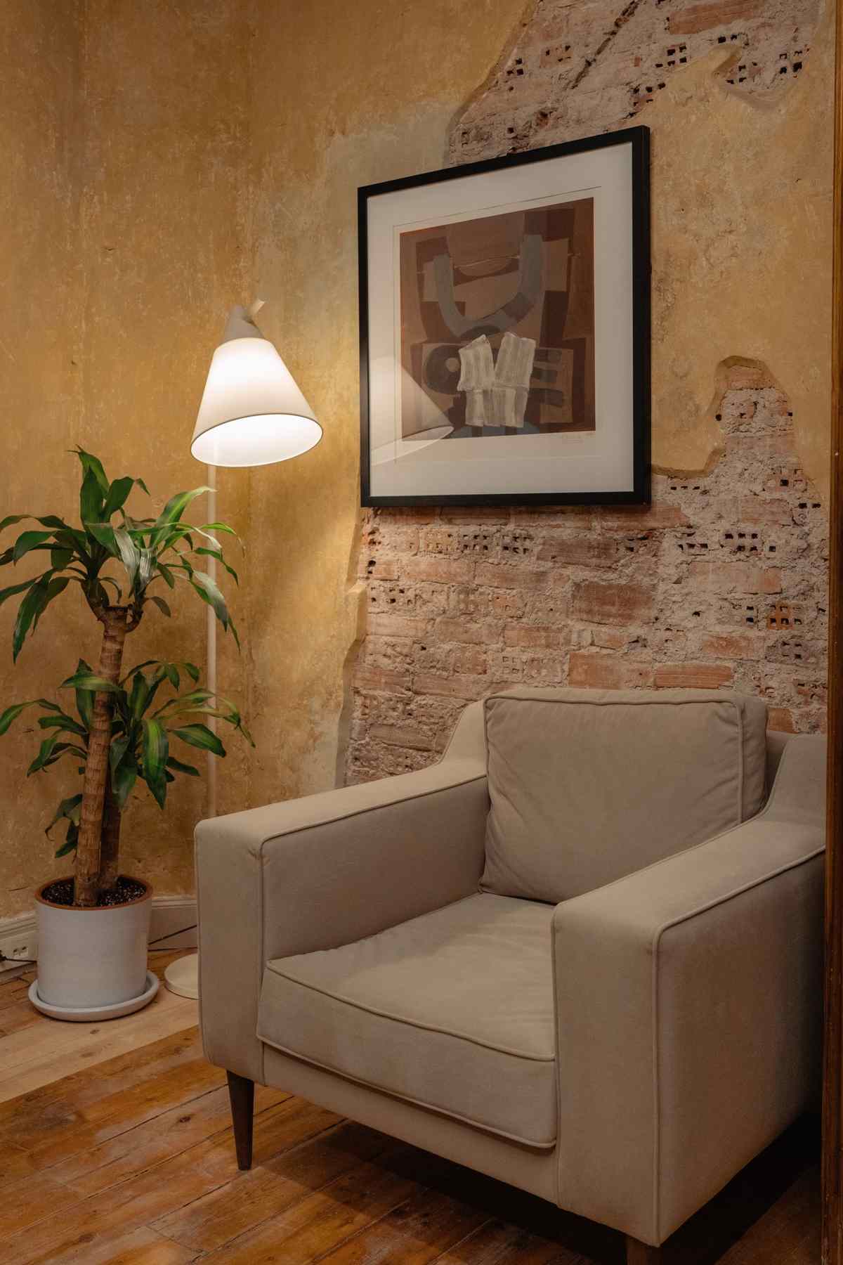 A framed abstract print on a textured wall above an armchair, lit by a white floor lamp beside a potted plant