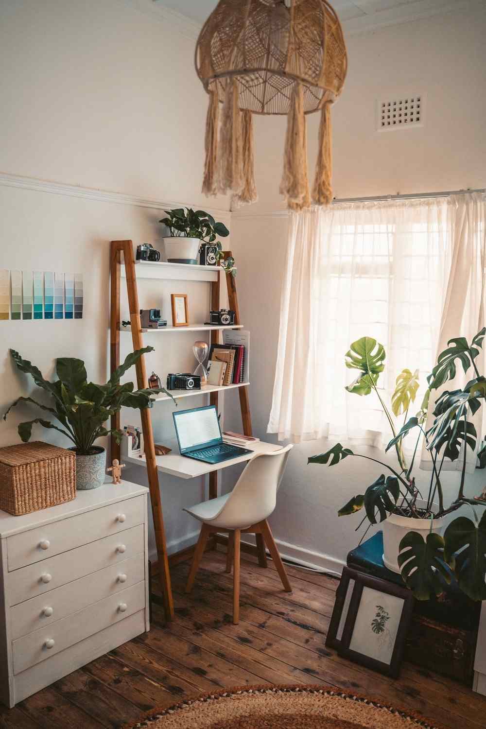 A home office corner with a ladder desk and laptop by a bright window, shelves holding cameras and a small framed print, and framed pictures leaning nearby