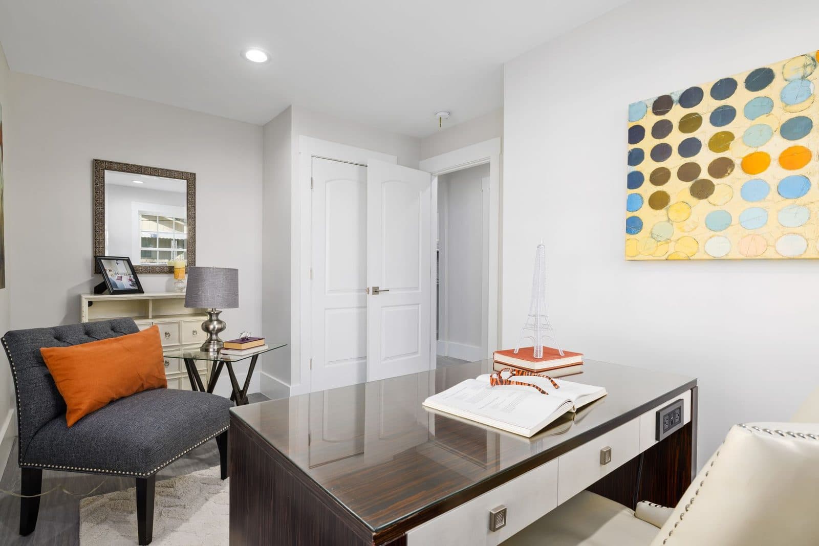 Bright home office with a dark wood desk, a grey chair with an orange cushion, and a colorful dot painting on the wall.