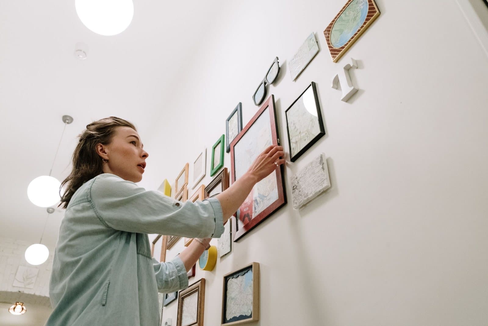 A woman adjusting a framed map print on a densely hung gallery wall of mixed frames, small artworks, and decorative objects.