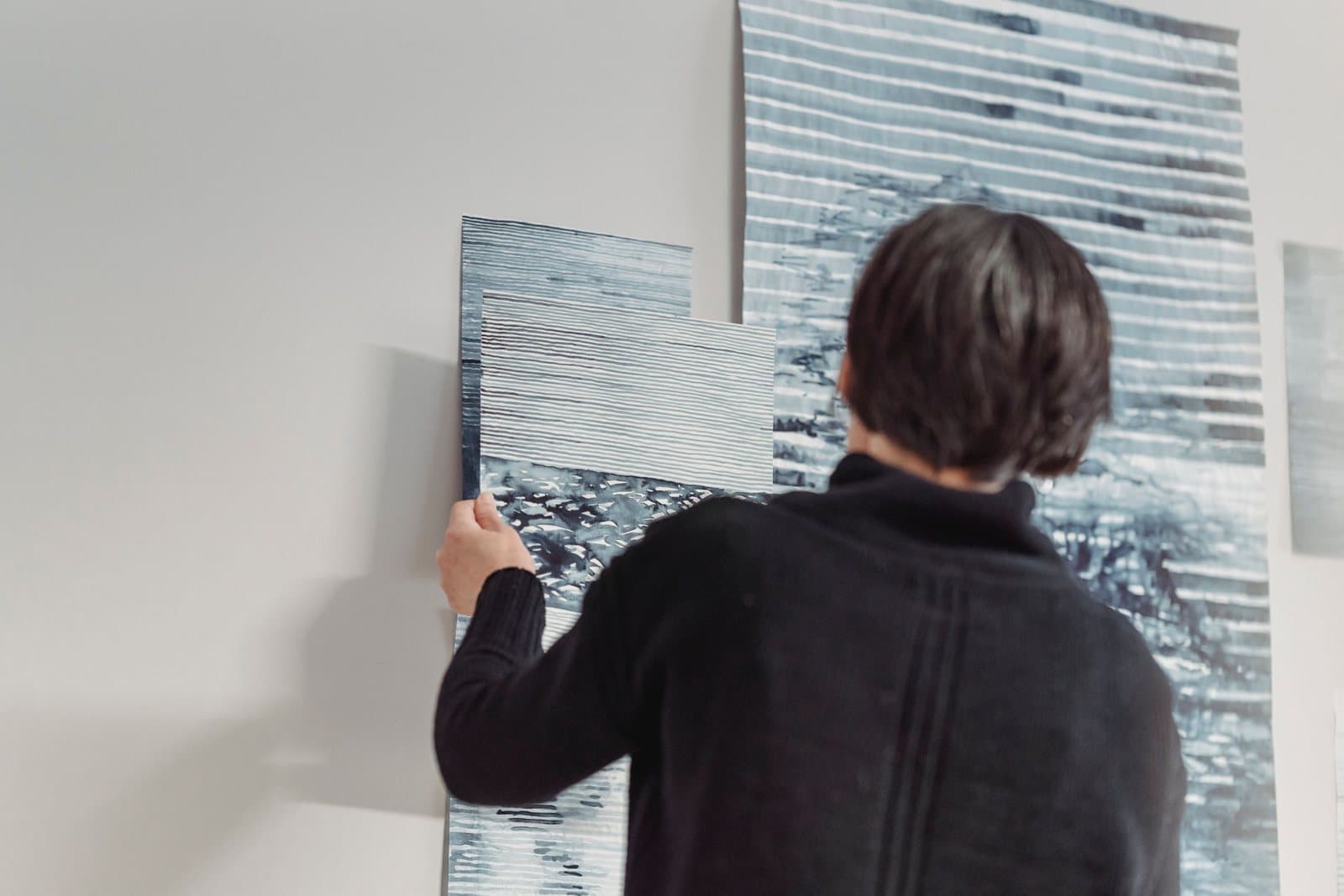 A person holding a large abstract canvas panel against a wall while positioning it beside a second oversized panel already in place.