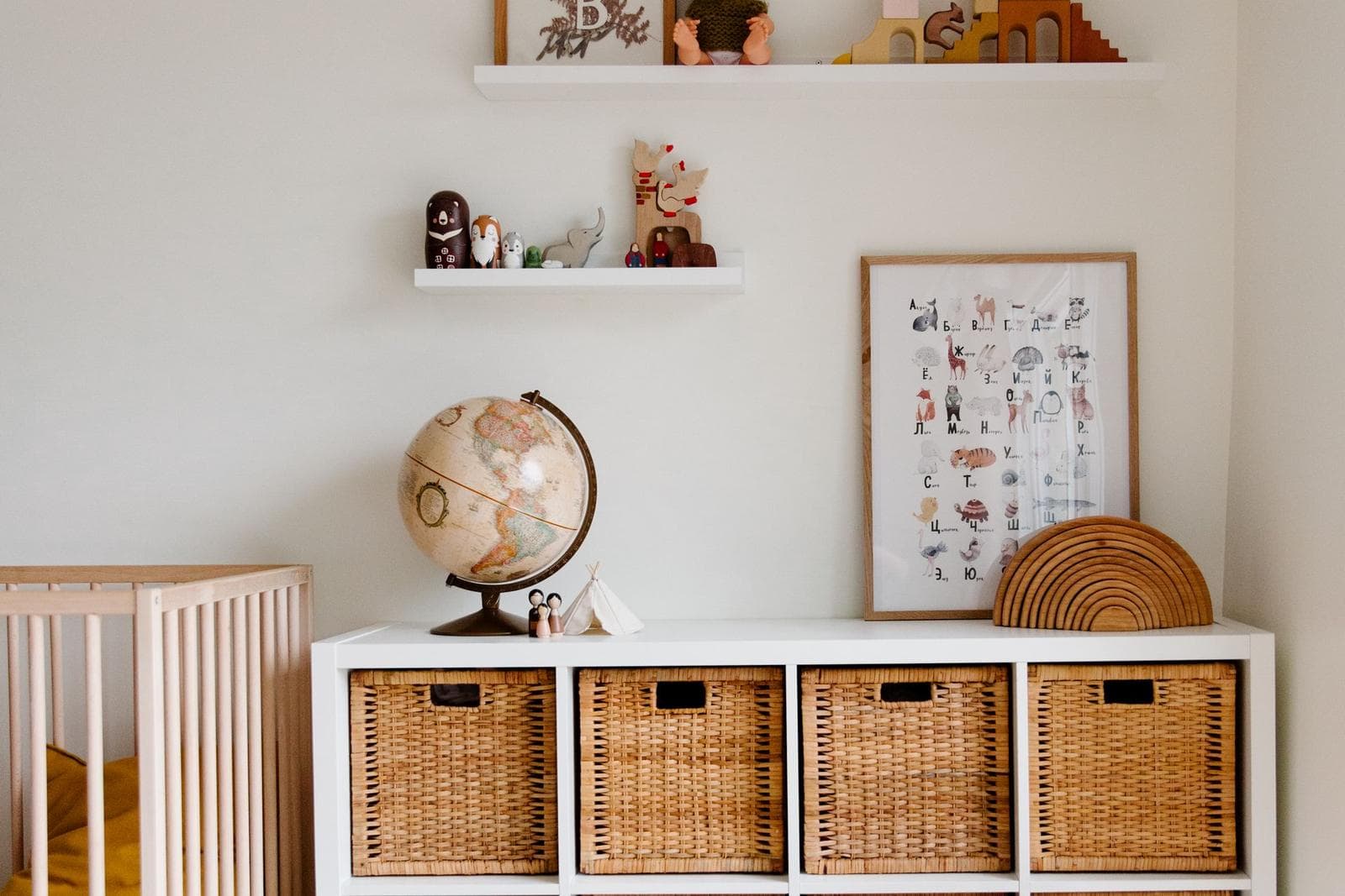 Child's bedroom corner with a globe and small figurines on white shelves above a storage unit with wicker baskets and a leaning framed poster.