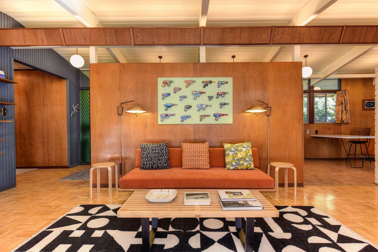 A mid-century modern living room with warm wood paneling, an orange sofa and a pop-art panel of colorful water pistols mounted above it