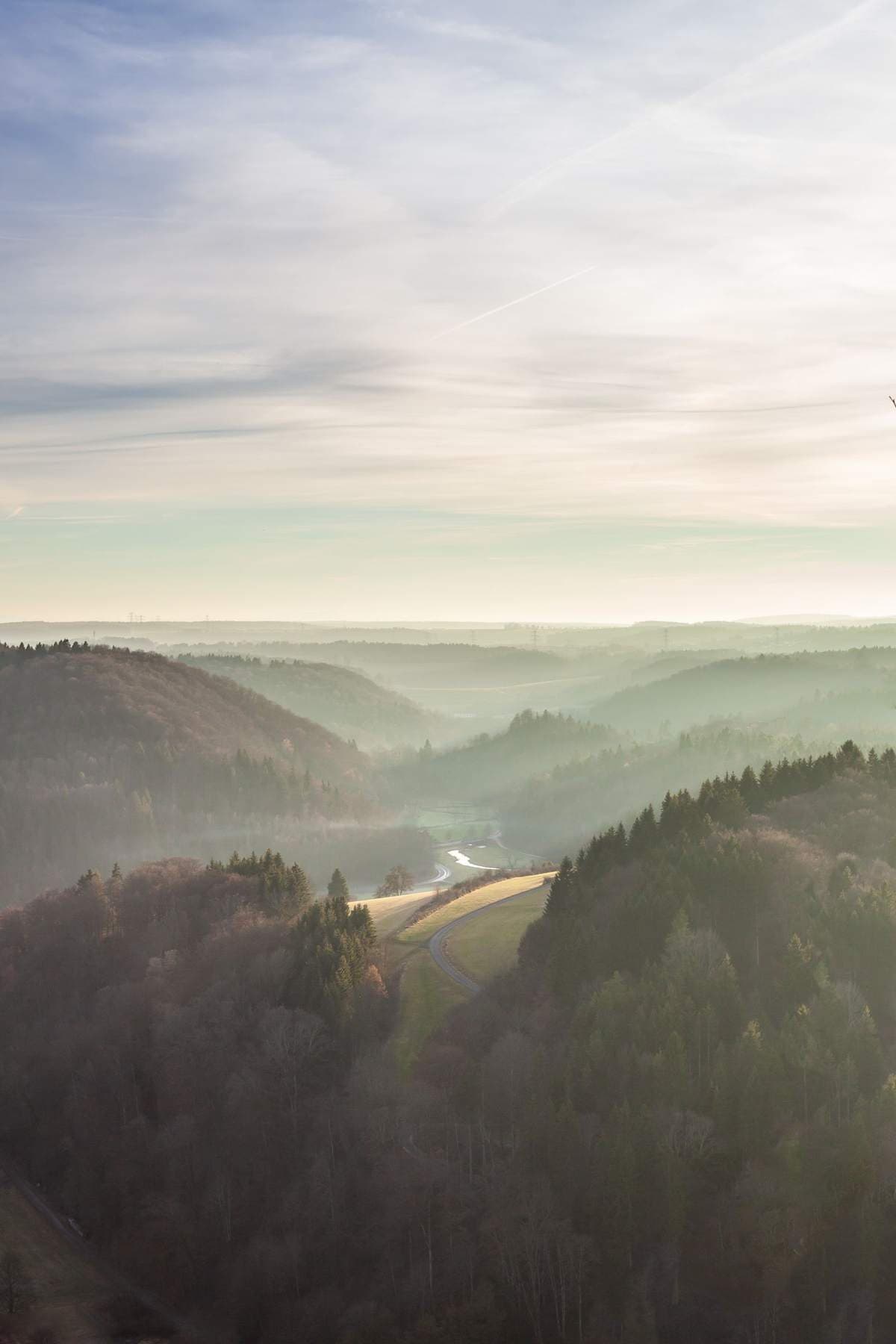 Layered misty hills and forested ridges receding into the distance above a winding road and river in a valley