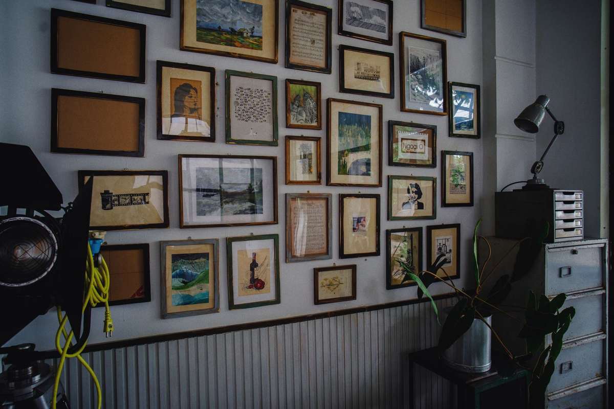 A wall covered with many framed paintings, prints and photographs in mixed wooden frames above a metal cabinet and a plant