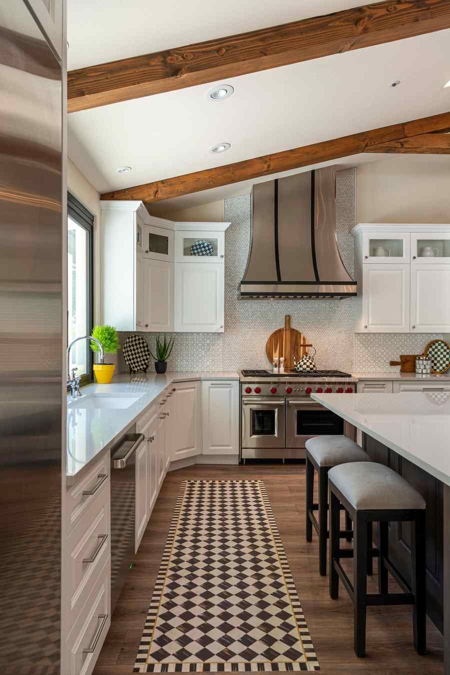 A modern farmhouse kitchen with white cabinets, wooden ceiling beams, a steel range hood and a patterned runner
