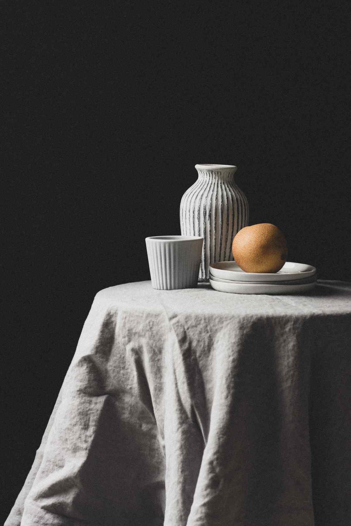 Minimalist still life of a pear on stacked plates beside a ribbed ceramic vase and cup on a draped table against a dark background