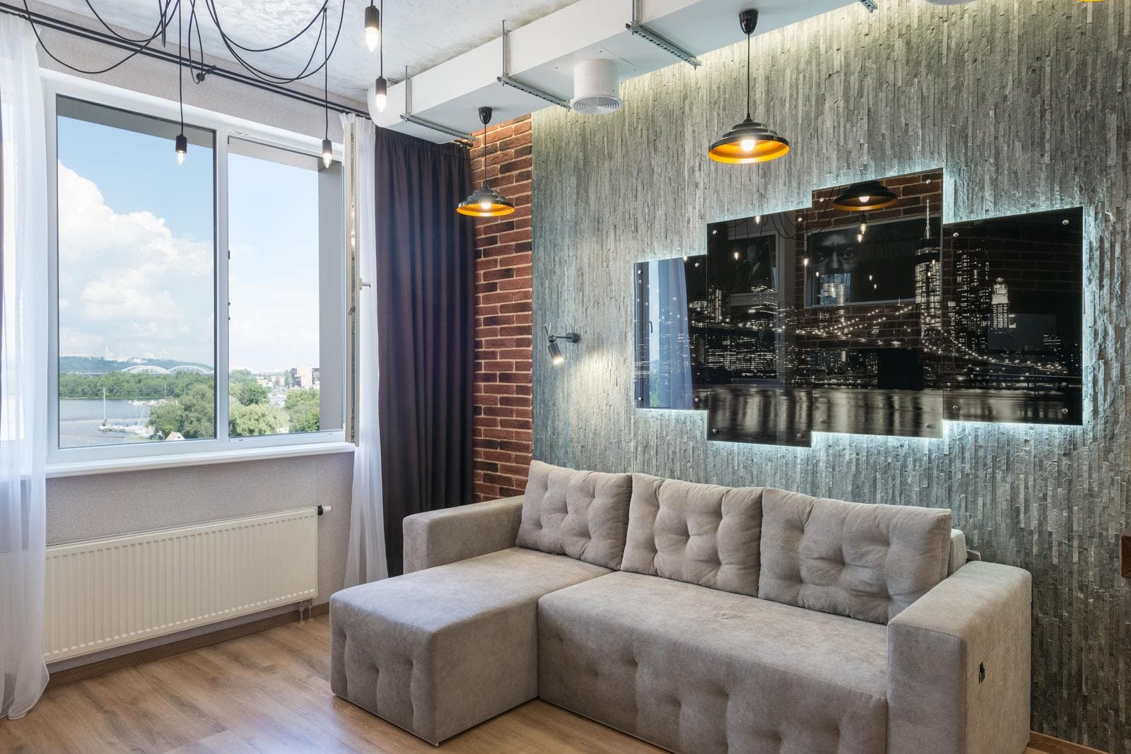 A five-panel split canvas print of a city skyline at night mounted on a textured wall above an L-shaped sofa in a loft-style living room.