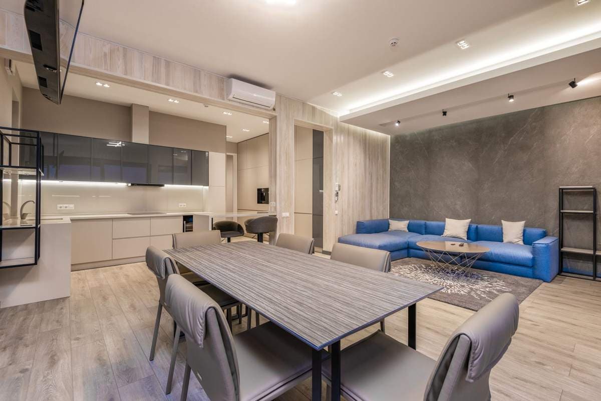 Open-plan apartment with a dining table and chairs in the foreground, a kitchen to one side, and a blue sofa living zone beyond