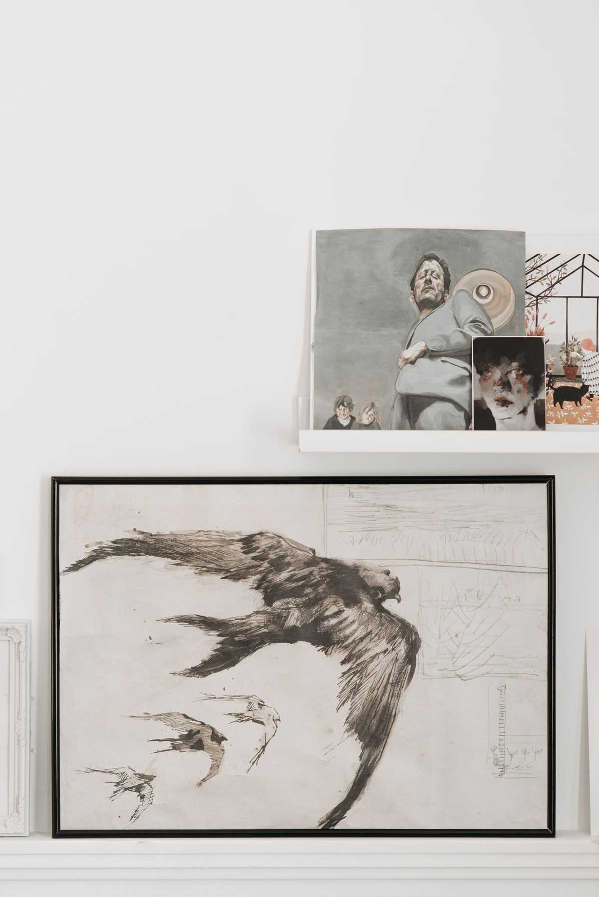 Framed and unframed paintings standing upright on white wall shelves, including a large ink drawing of a bird in flight