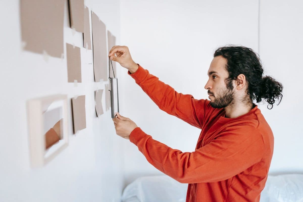 A man holding a small frame by its chain in front of a white wall marked out with taped paper templates