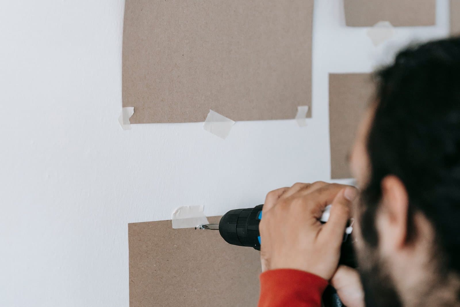 A man driving a screw into a wall with a drill-driver through the marked corner of a taped paper frame template.