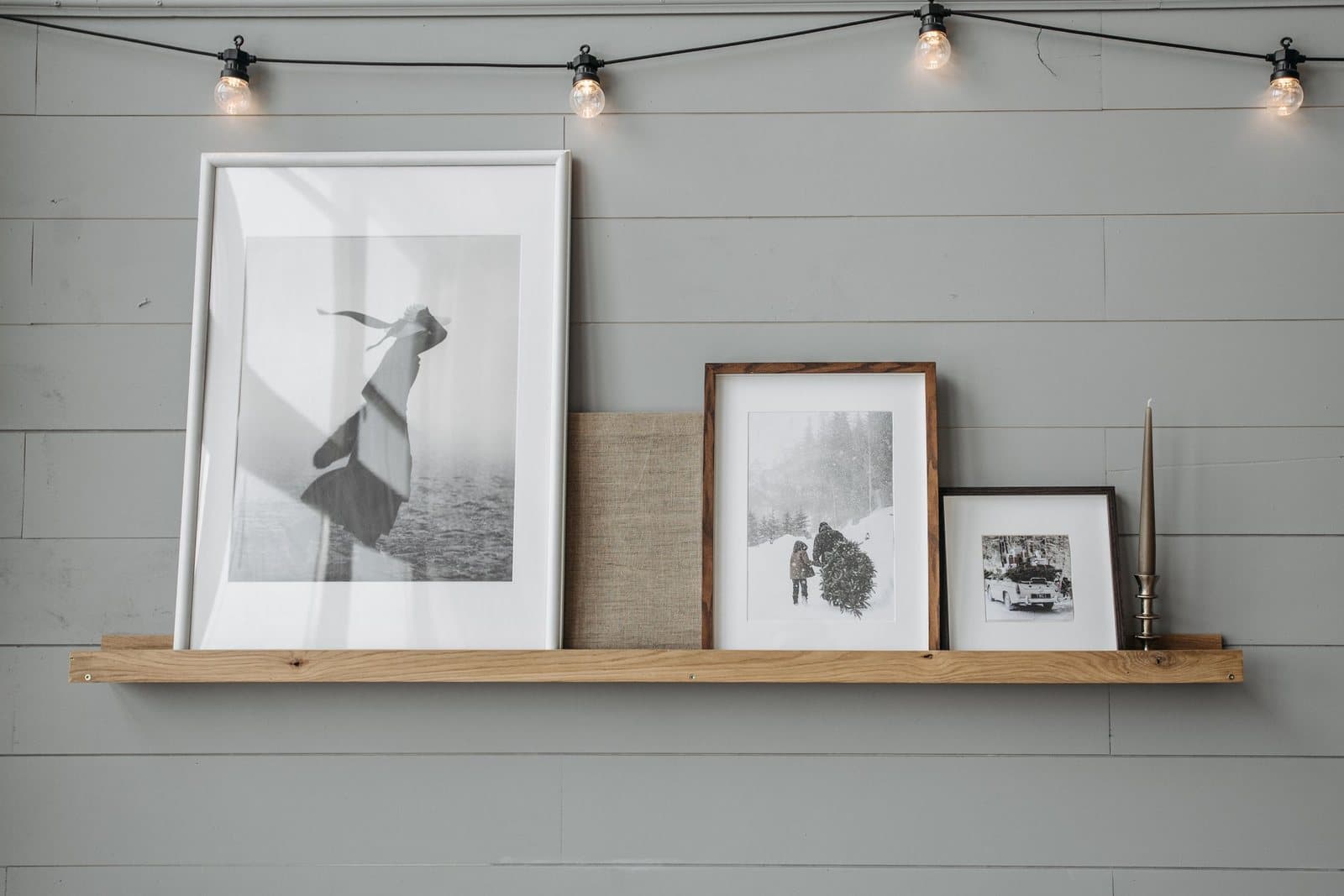 Layered framed black-and-white photographs and a tall candle arranged along a wooden picture ledge on a gray shiplap wall.