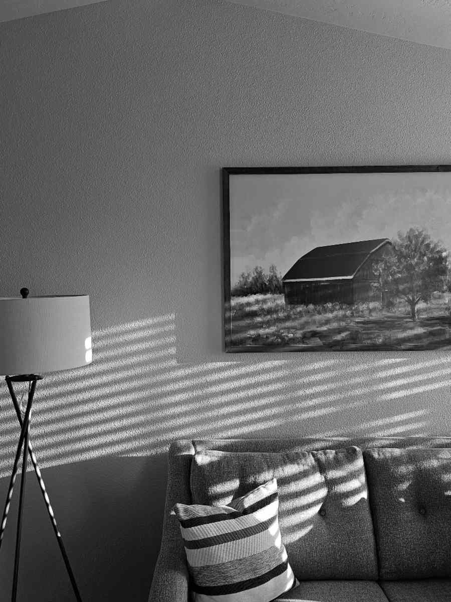 Black-and-white photograph of a large framed barn painting hanging alone above a sofa beside a floor lamp