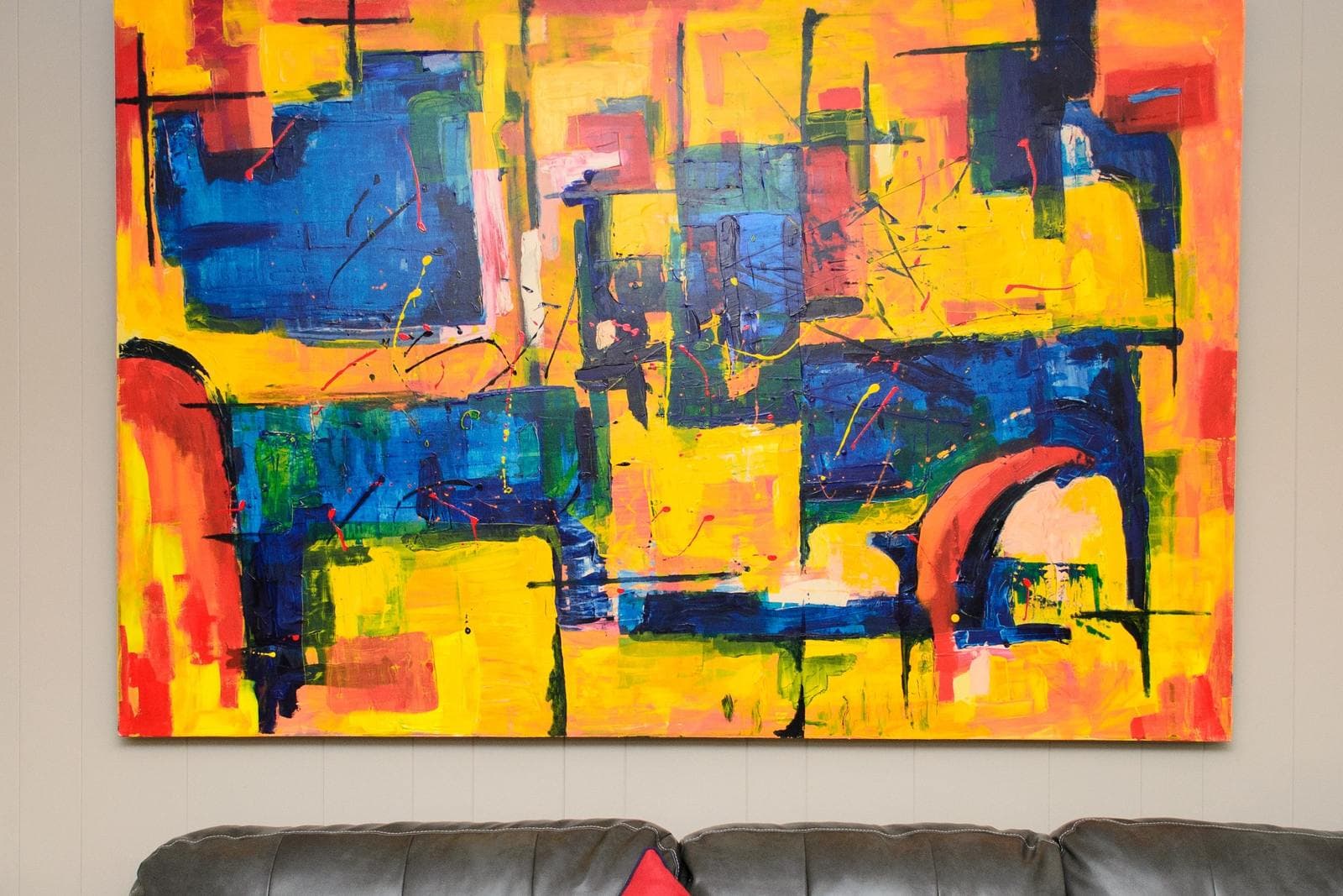 A single large, vividly colored abstract canvas in yellow, blue and red dominating the wall above a dark leather sofa.