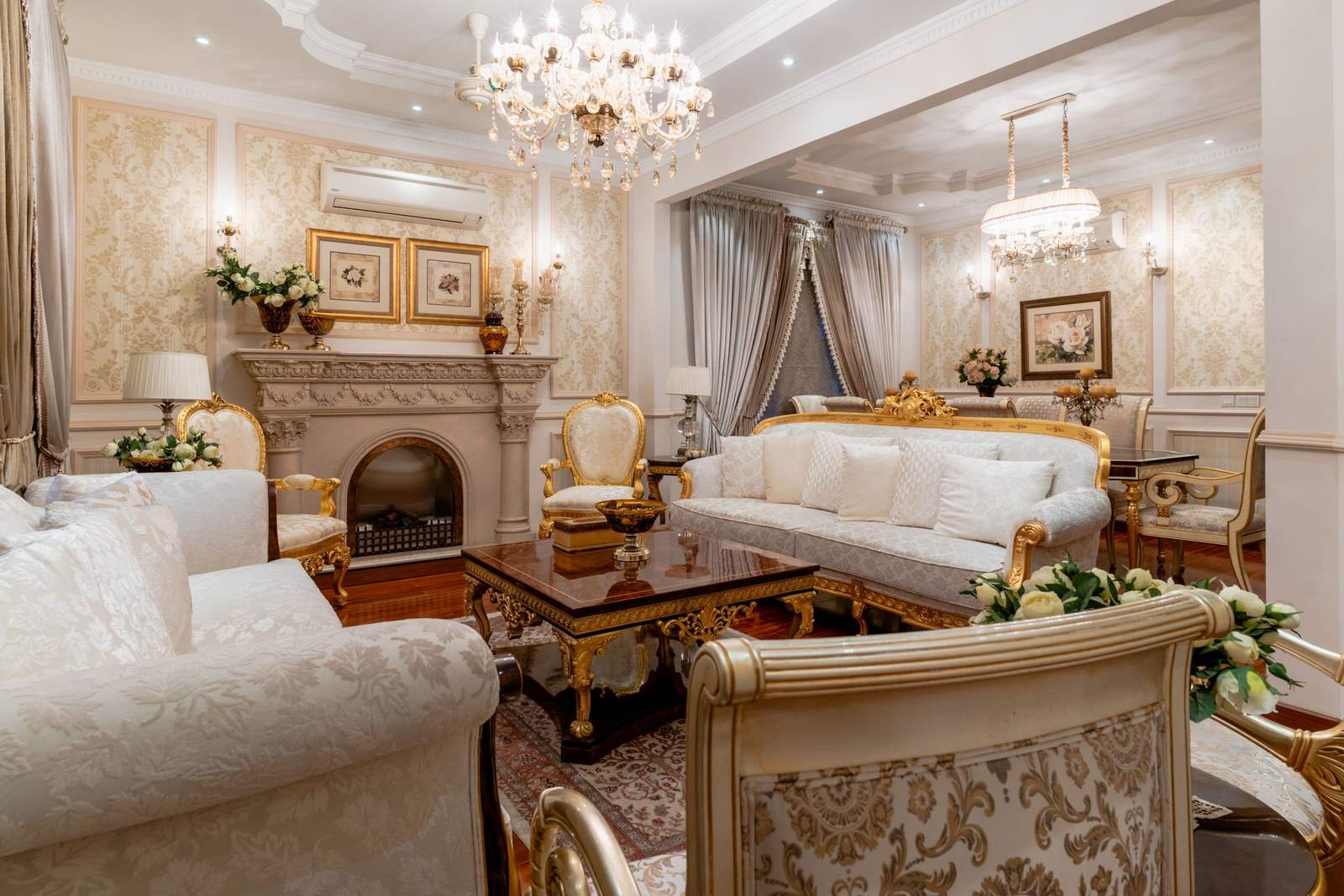 A formal traditional living room with damask wallpaper, crystal chandeliers and a pair of gilt-framed prints hanging above an ornate fireplace